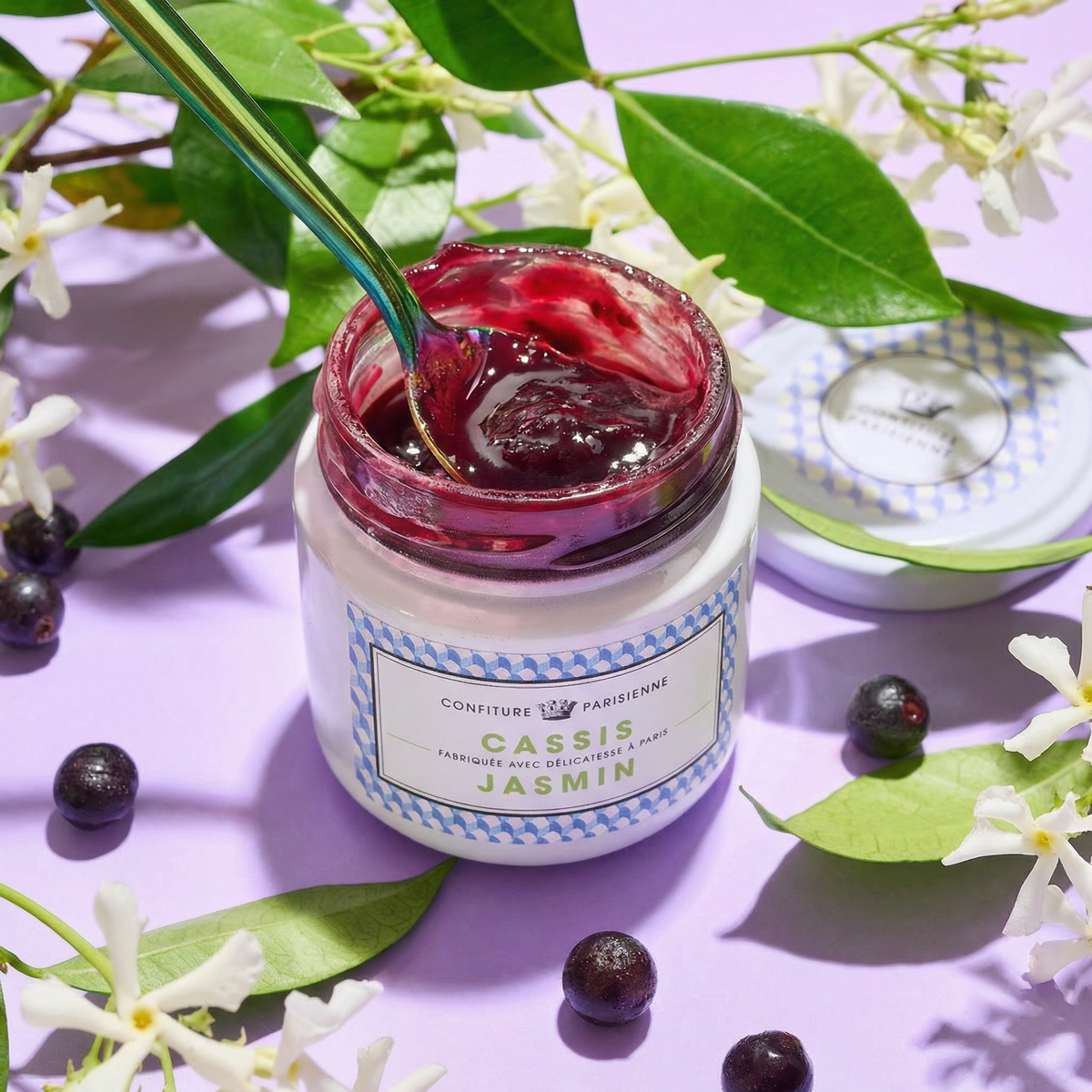 Pot ouvert de confiture Cassis Jasmin sur fond lilas, entouré de fleurs de jasmin et de baies de cassis
