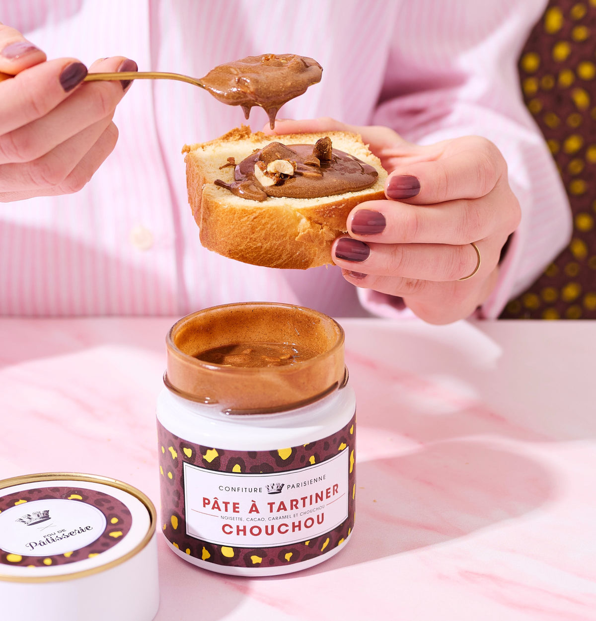 Mains tartinant une tranche de brioche de Pâte à tartiner Chouchou à la cuillère, pot ouvert au premier plan