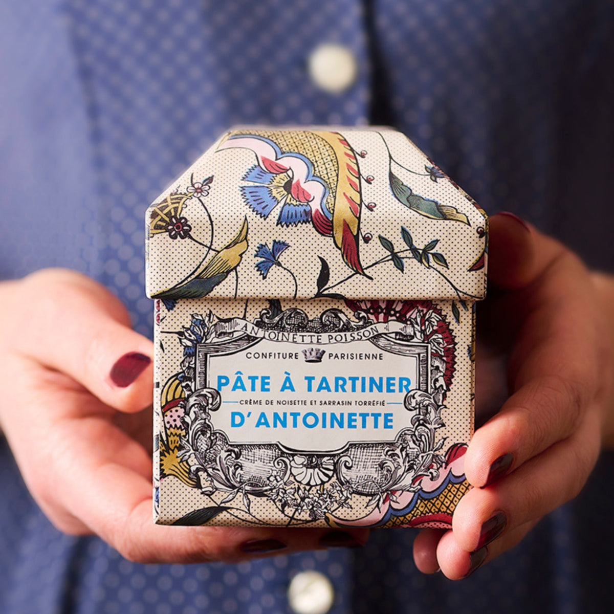 Mains tenant le coffret fleuri de la Pâte à Tartiner d'Antoinette x Antoinette Poisson devant une chemise bleue