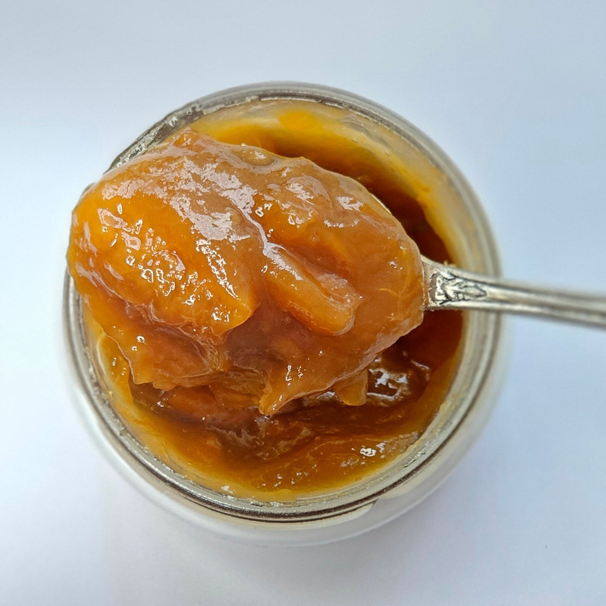 Cuillère en argent chargée d'une confiture orangée avec morceaux de fruit au-dessus d'un pot ouvert, vue de dessus