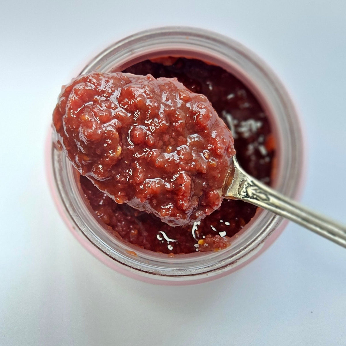 Cuillère en argent chargée de confiture de fraise en morceaux au-dessus d'un pot ouvert, vue de dessus