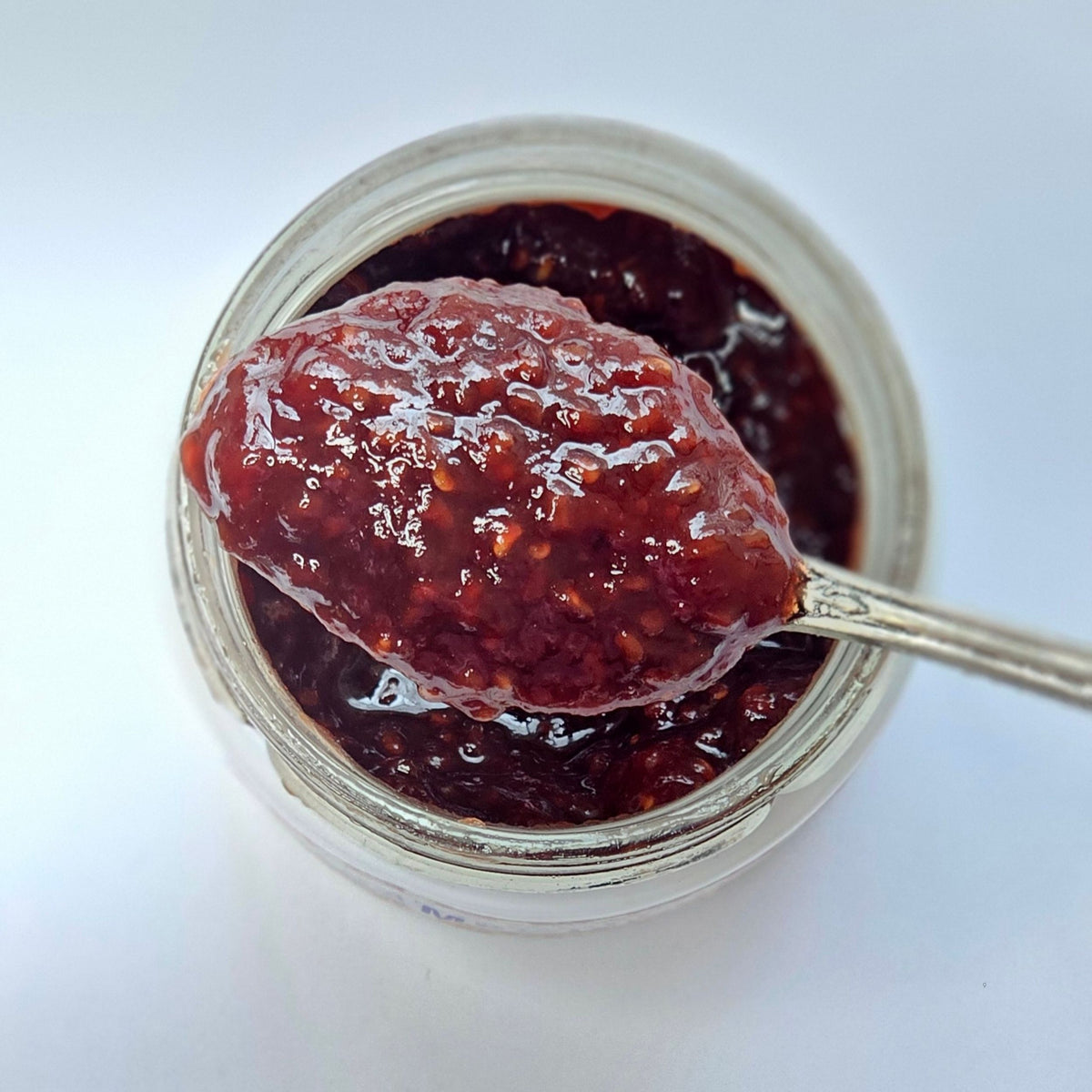 Cuillère en argent chargée de confiture rouge sombre à grains au-dessus d'un pot ouvert, vue de dessus