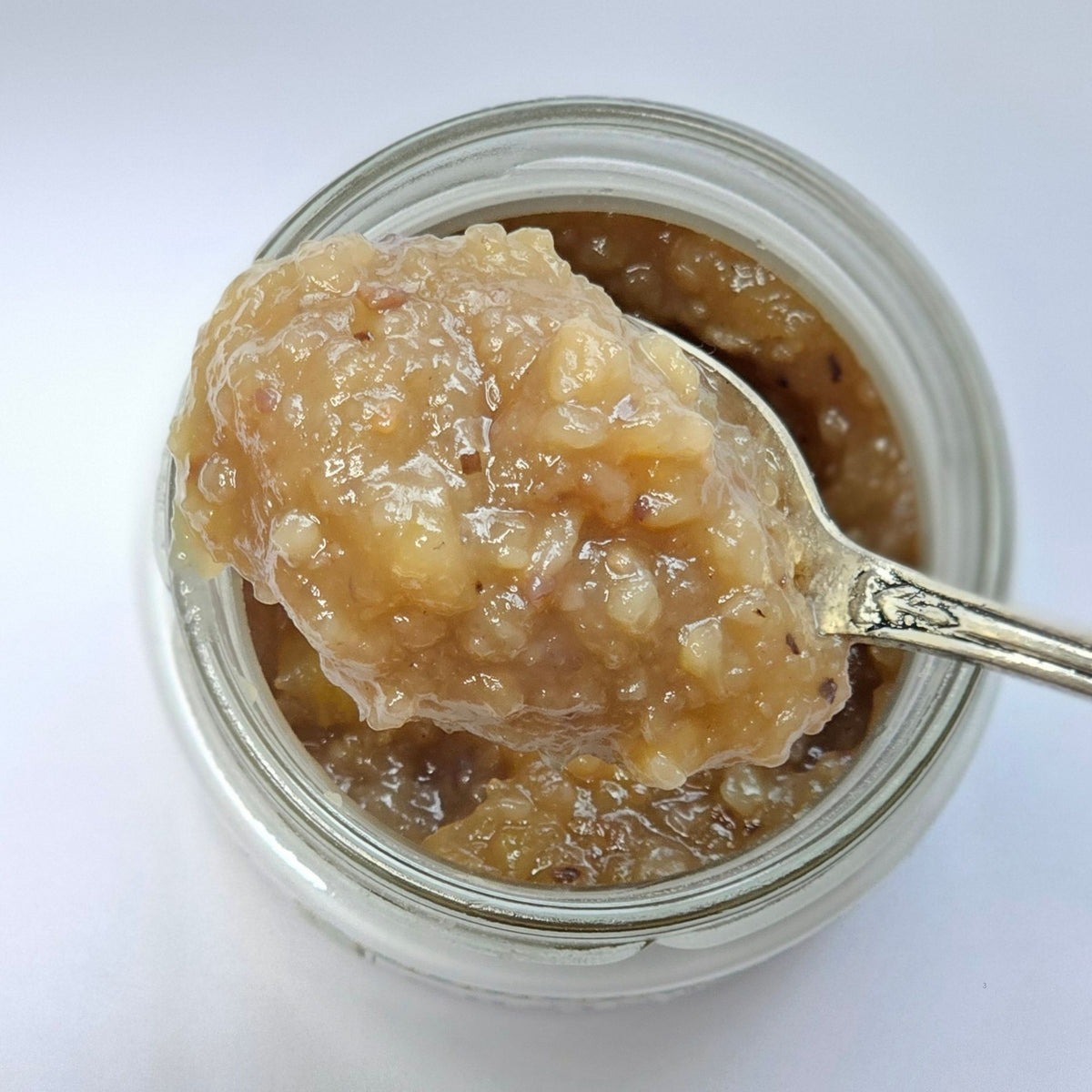 Cuillère en argent chargée d'une confiture claire en morceaux, type poire ou pomme, au-dessus d'un pot ouvert