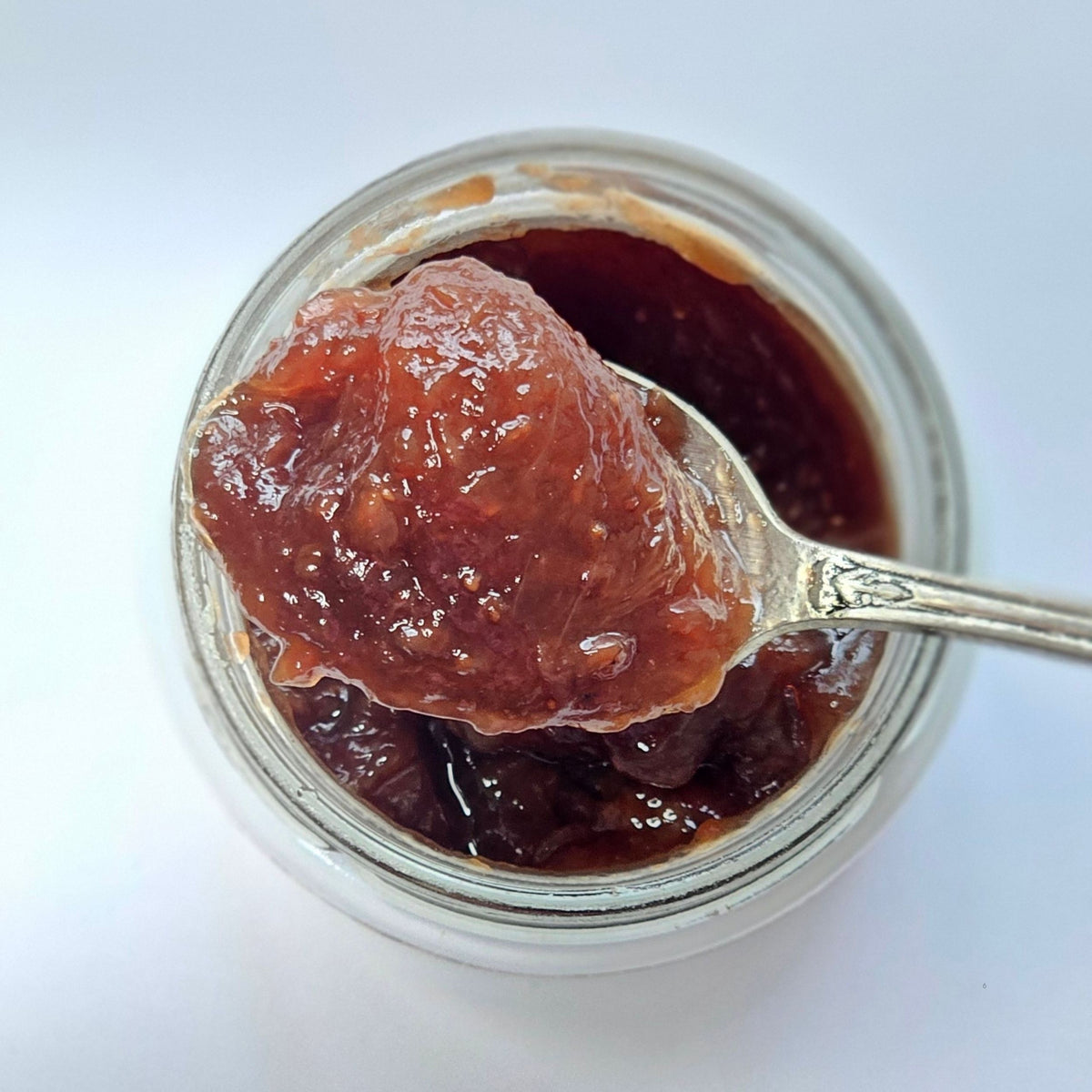 Cuillère en argent chargée de confiture rouge brun au-dessus d'un pot ouvert, vue de dessus