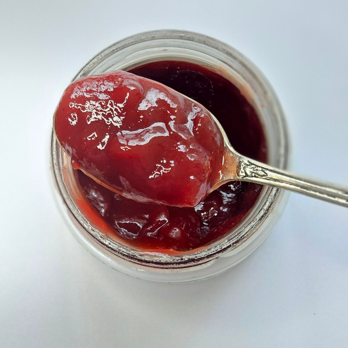 Cuillère en argent chargée de confiture rouge brillante au-dessus d'un pot ouvert, vue de dessus