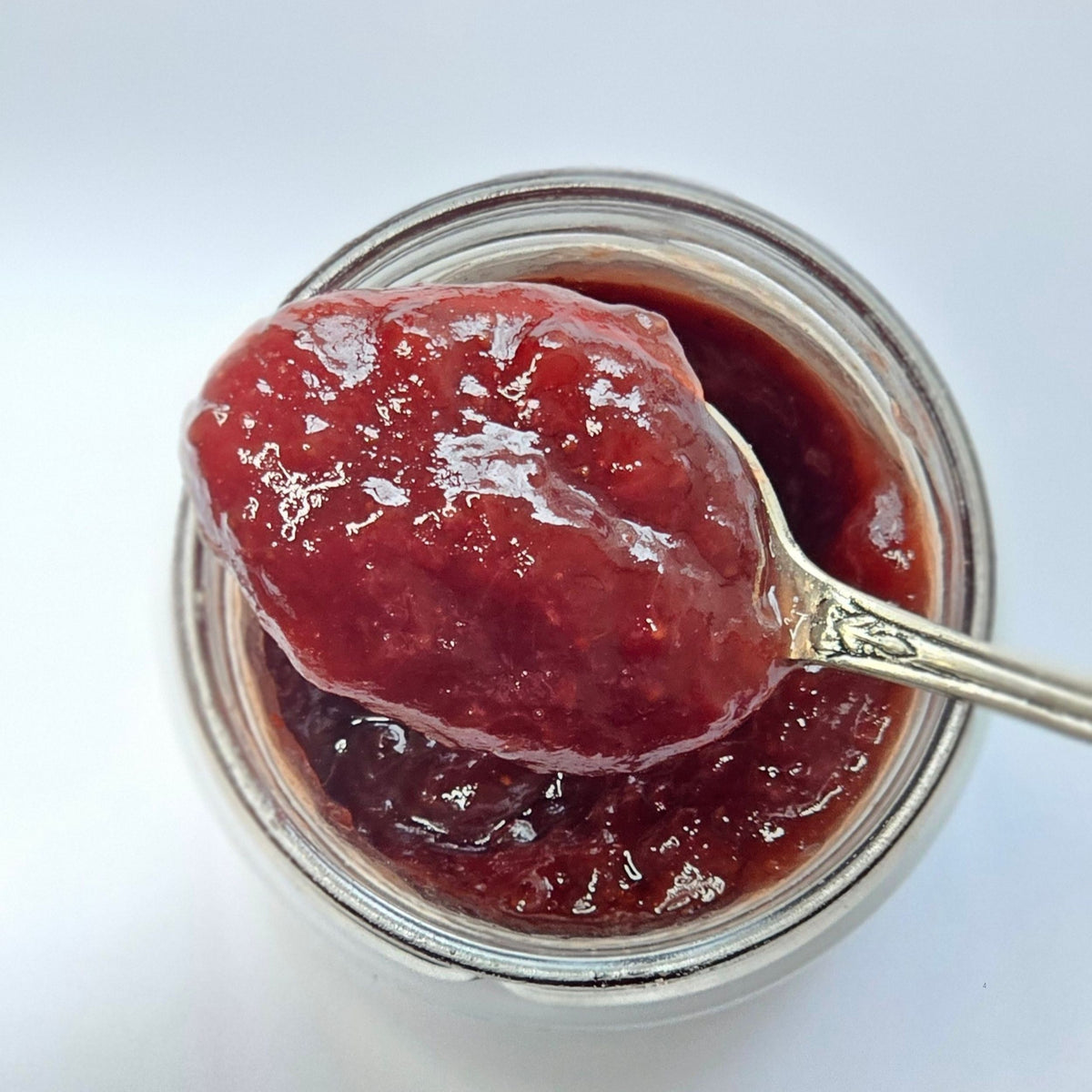 Cuillère en argent chargée de confiture rouge au-dessus d'un pot ouvert, vue de dessus