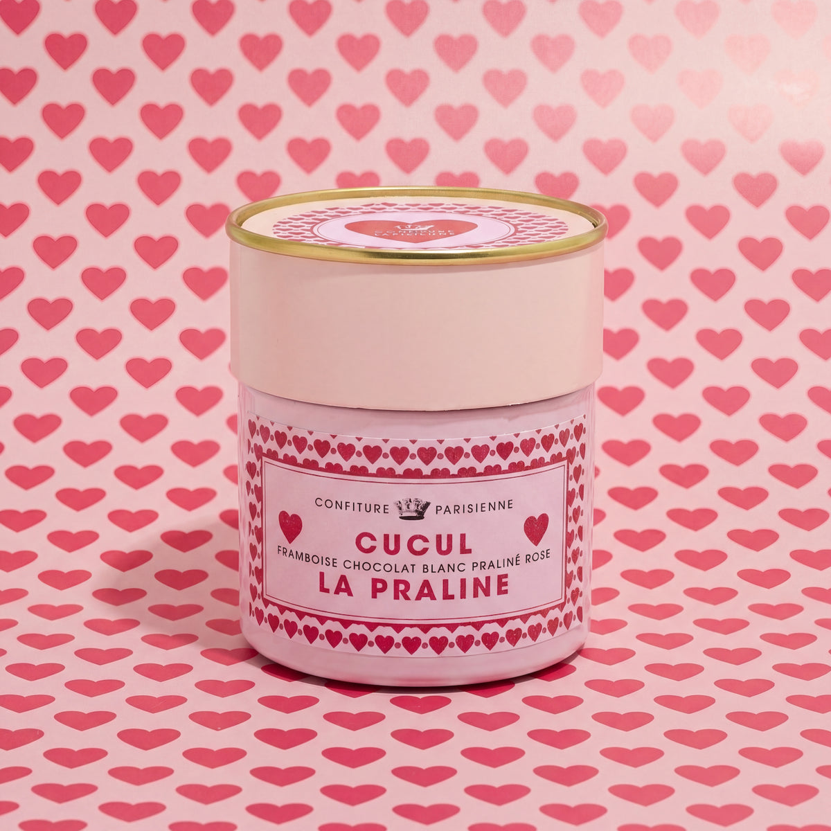 Pot Cucul la Praline posé devant un fond rose imprimé de petits cœurs rouges