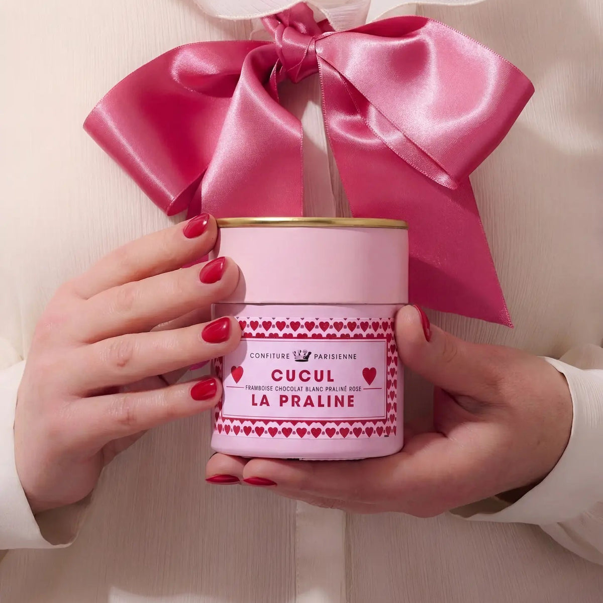 Image générée : mains aux ongles rouges tenant un pot Cucul la Praline orné d'un grand nœud en satin rose