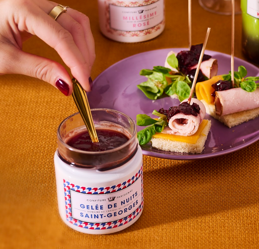 Pot ouvert de Gelée de Nuits-Saint-Georges avec une cuillère dorée, à côté d'une assiette de bouchées apéritives