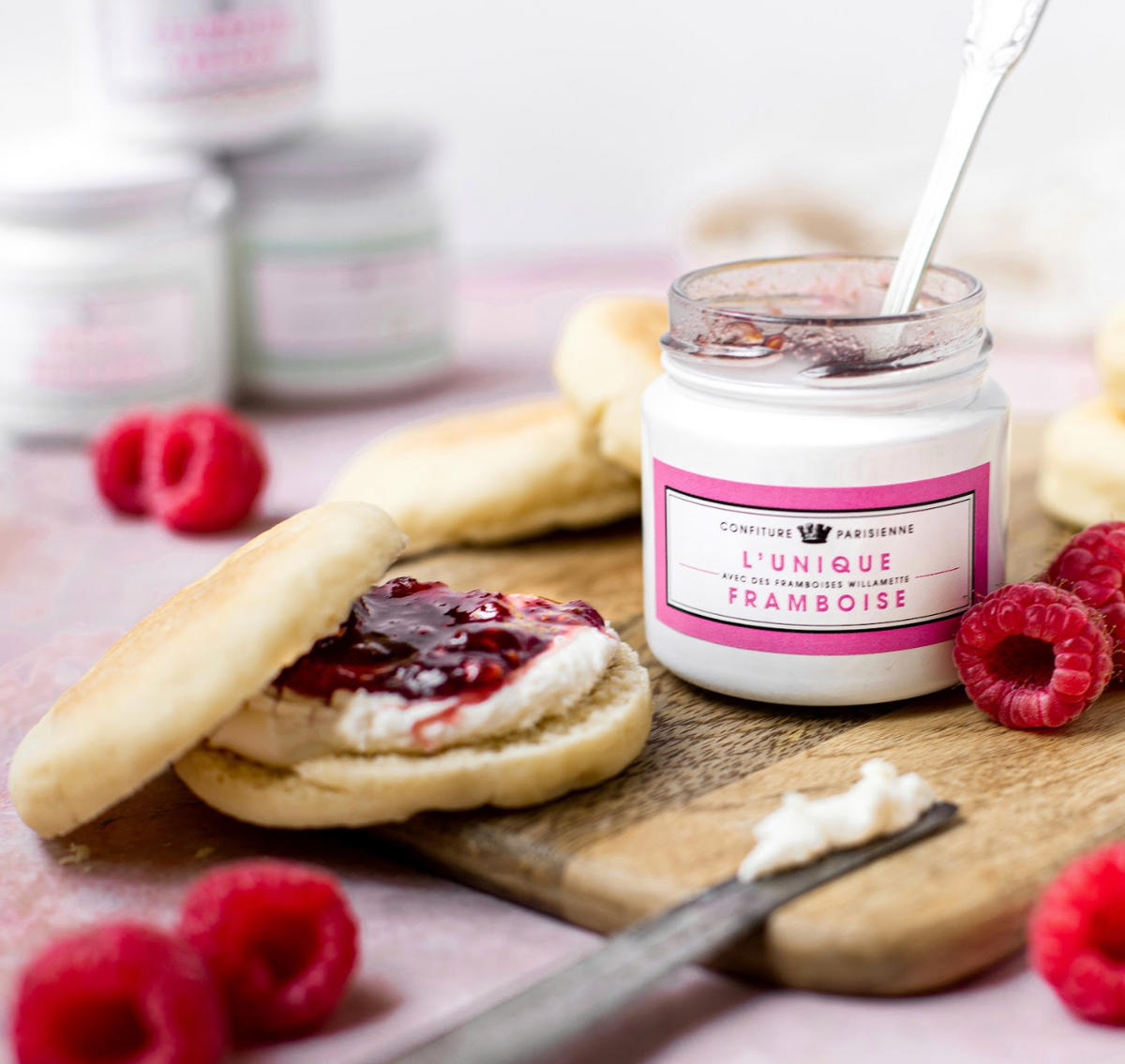 Pot de L'Unique Framboise avec des crumpets tartinés de fromage frais et de confiture, framboises fraîches autour