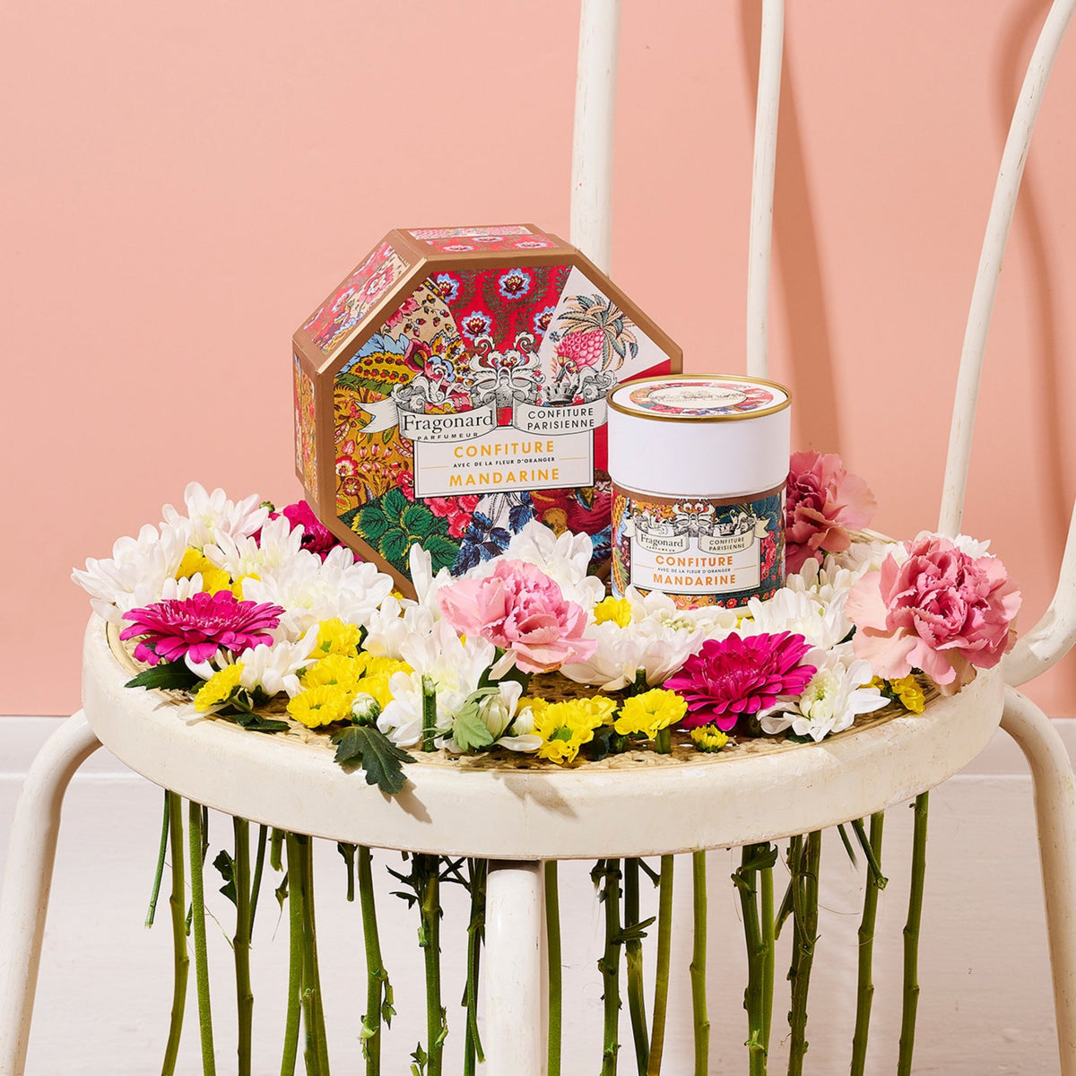 Coffret et pot Mandarine Fleur d'Oranger x Fragonard sur une chaise blanche garnie de fleurs, fond rose