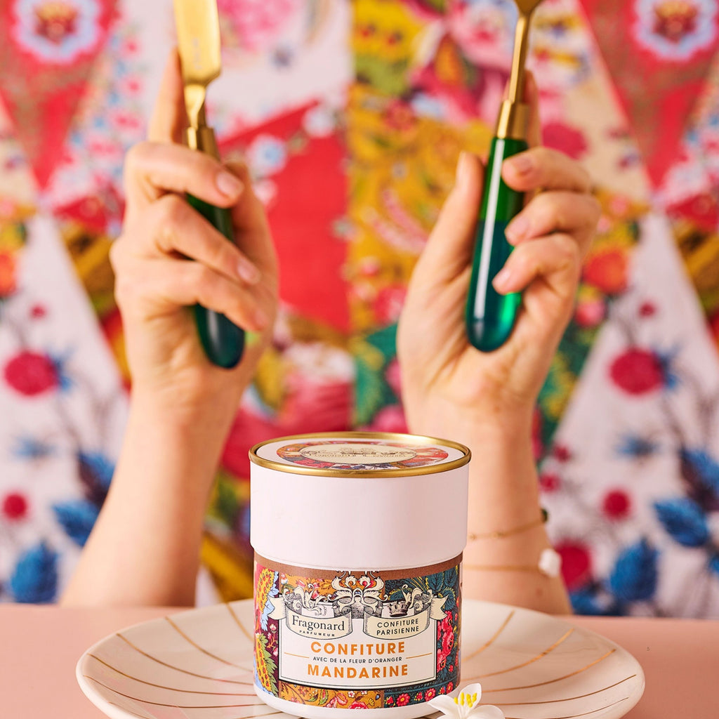 Pot de Confiture Mandarine Fleur d'Oranger x Fragonard sur une assiette, deux mains tenant des couverts verts devant un tissu fleuri