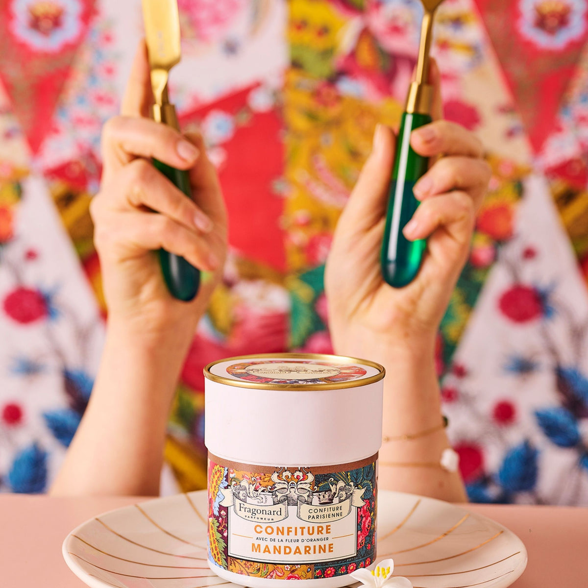 Pot de Confiture Mandarine Fleur d'Oranger x Fragonard sur une assiette, deux mains tenant des couverts verts devant un tissu fleuri