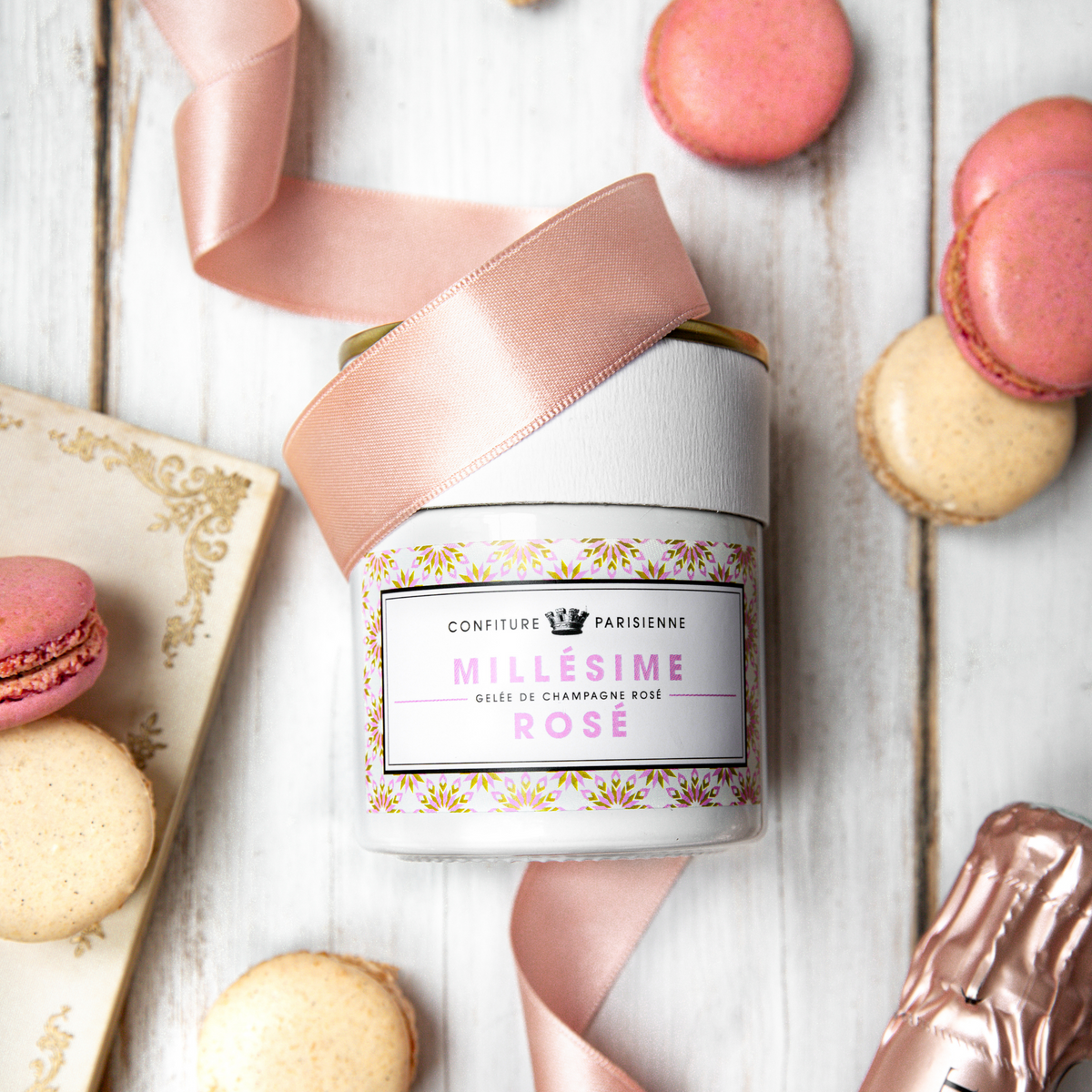 Pot de Gelée Millésime Champagne Rosé couché sur une table blanche avec des macarons et un ruban rose